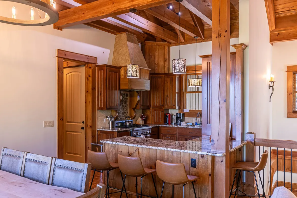 The image spotlights a gourmet kitchen with a substantial island, high-end appliances (including Thermador), wooden beams overhead, bar stools for casual dining, and elegant countertops. The kitchen is designed for both culinary adventure and social gathering, with a welcoming and open feel.