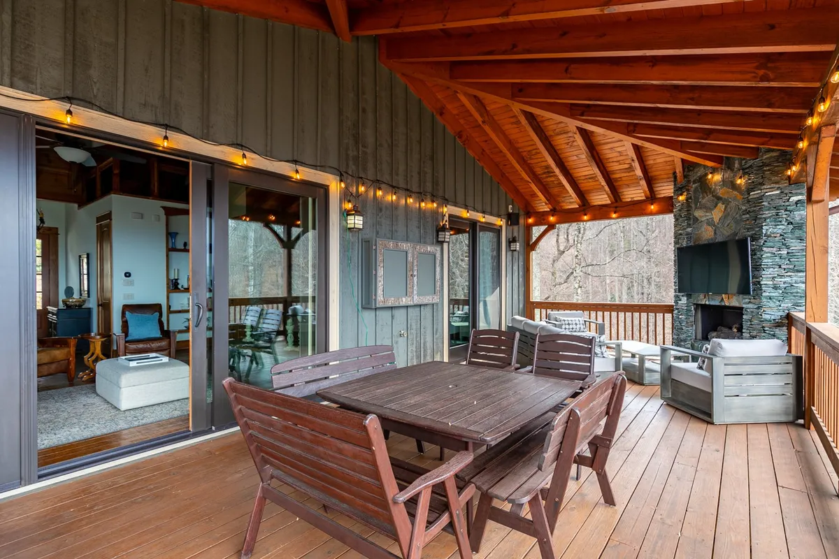 A covered outdoor living area featuring a large wooden dining table, a striking stone fireplace, comfortable lounge seating, and festive string lights overhead. The space exudes warmth and versatility, ideal for gatherings year-round—whether for summer barbecues or intimate winter evenings by the fire.