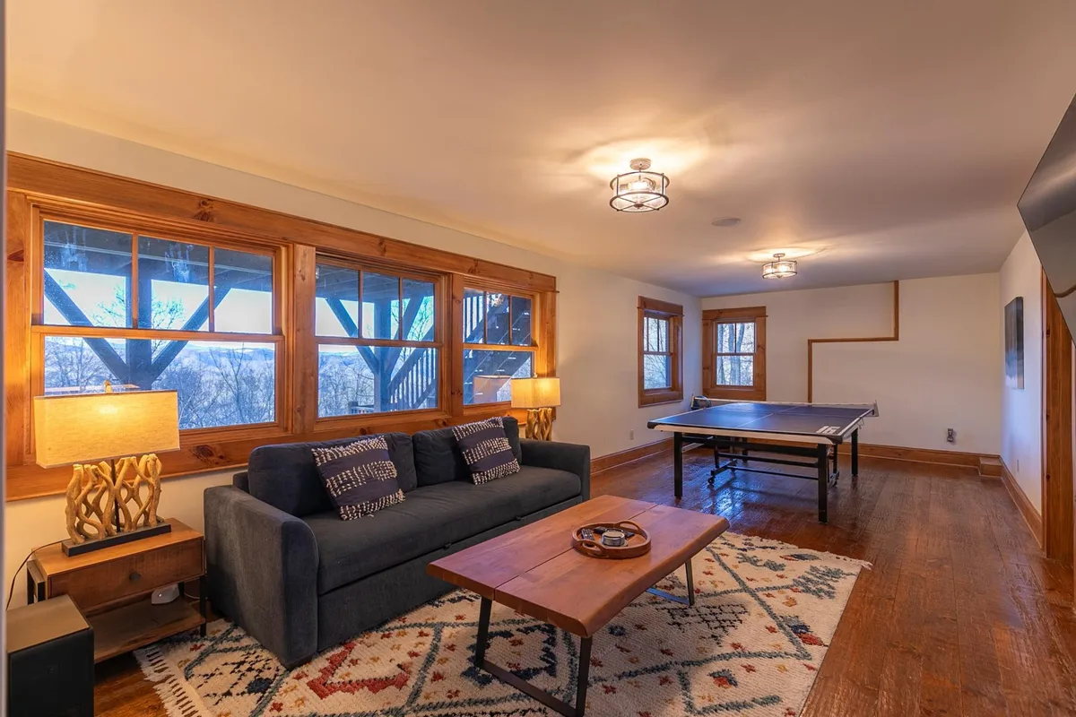 A multi-functional living space is depicted here, with a cozy sofa facing a ping pong table and a coffee table. Large windows let in natural light and offer a glimpse of the outdoors, while the arrangement encourages both play and conversation, mirroring the home's secondary living and game area setup.