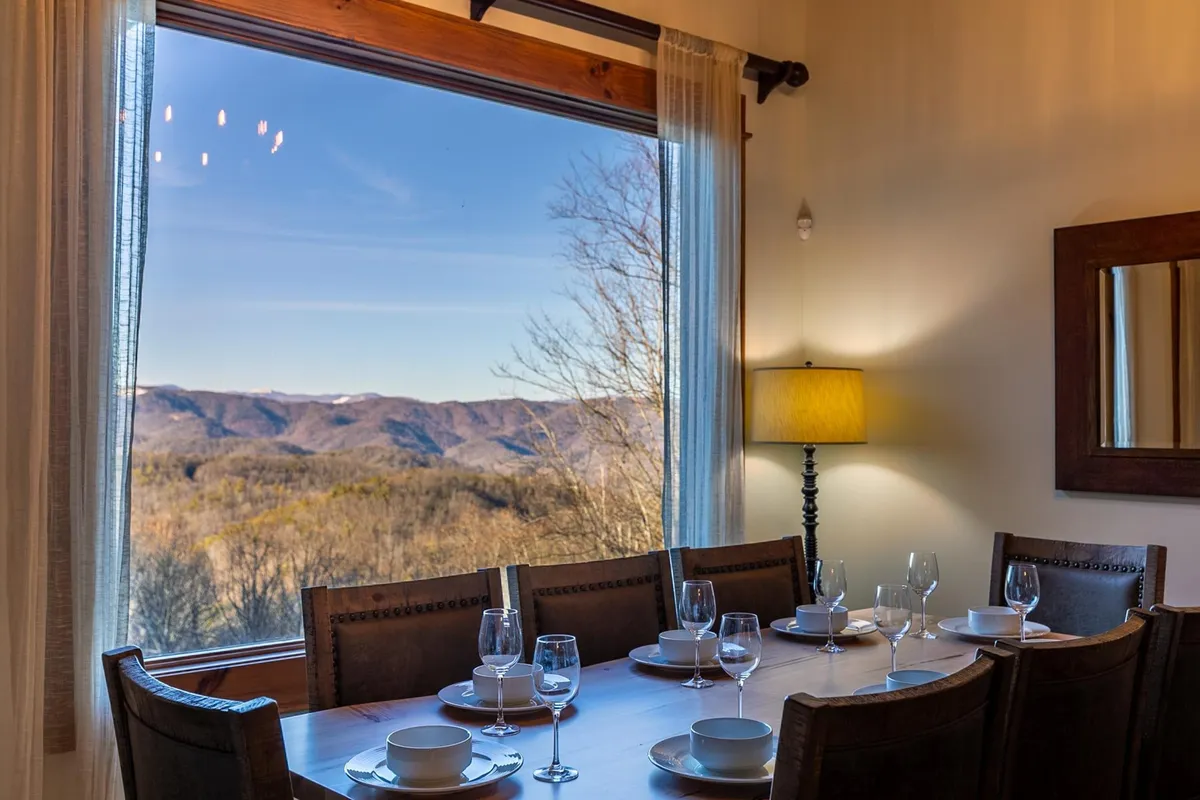 A large dining table is set for a meal, positioned beside a vast window offering a sweeping view of the Blue Ridge Mountains. The space is bathed in natural light, creating a welcoming spot for celebrations, family dinners, and memorable gatherings.