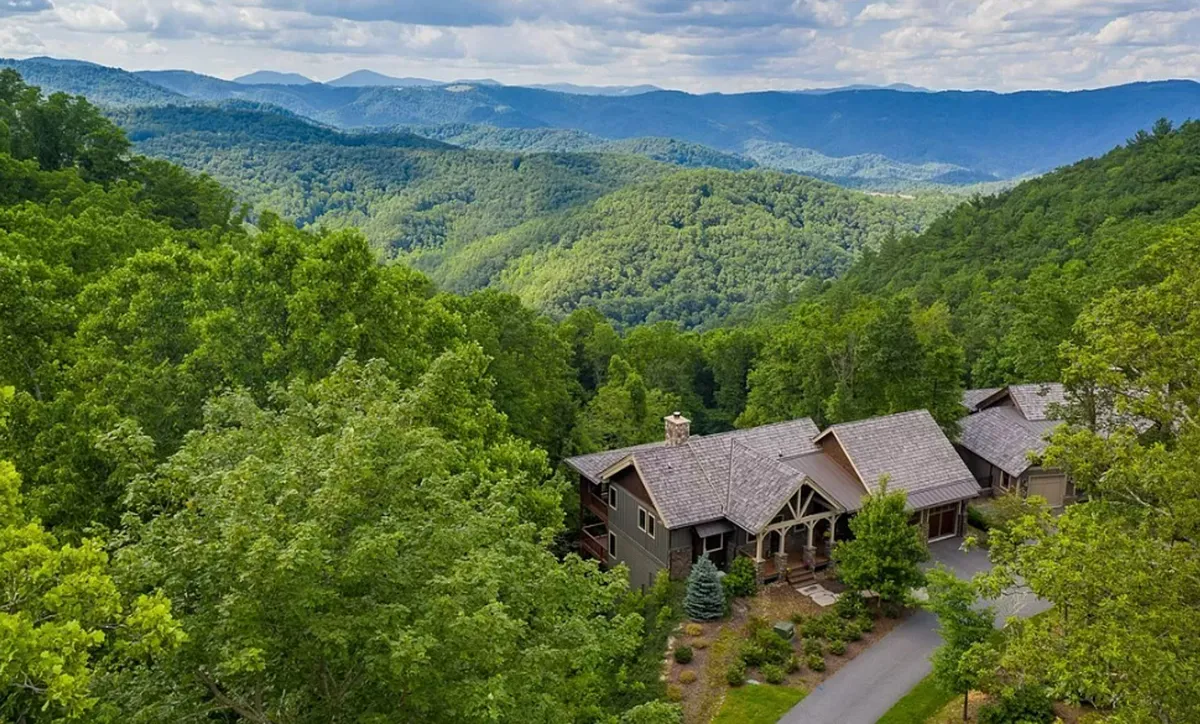 A tranquil exterior scene showing a panoramic view of the mountain range and dense wooded landscape, with a home nestled in the scenery. The setting evokes a sense of wilderness and direct access to nature, with the sprawling mountains and forest inviting exploration and adventure.
