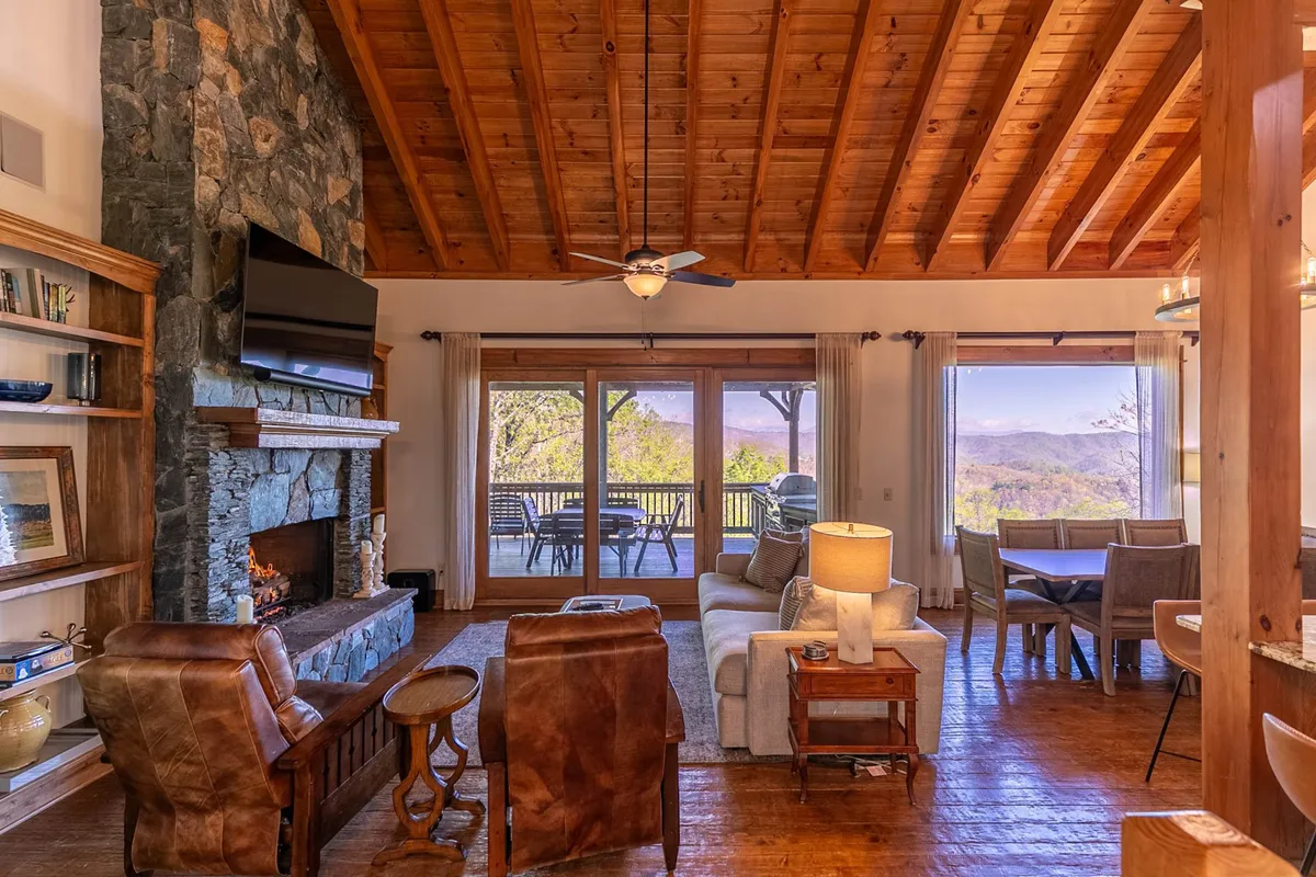 This image captures an open-concept living and dining area featuring a striking stone fireplace that reaches up to the ceiling, exposed wooden beams, large windows framing panoramic mountain views, and a comfortable seating arrangement. The dining table is positioned to enjoy scenic vistas, and the entire space exudes warmth and sociability.