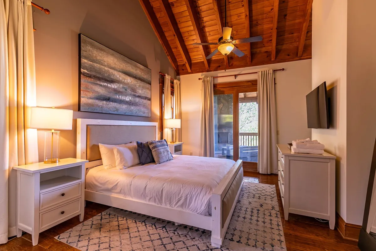 A serene bedroom with a large bed, tasteful artwork, nightstands, a window letting in natural light, and a wall-mounted television. The scene is calm and inviting, emphasizing restful comfort and relaxation.