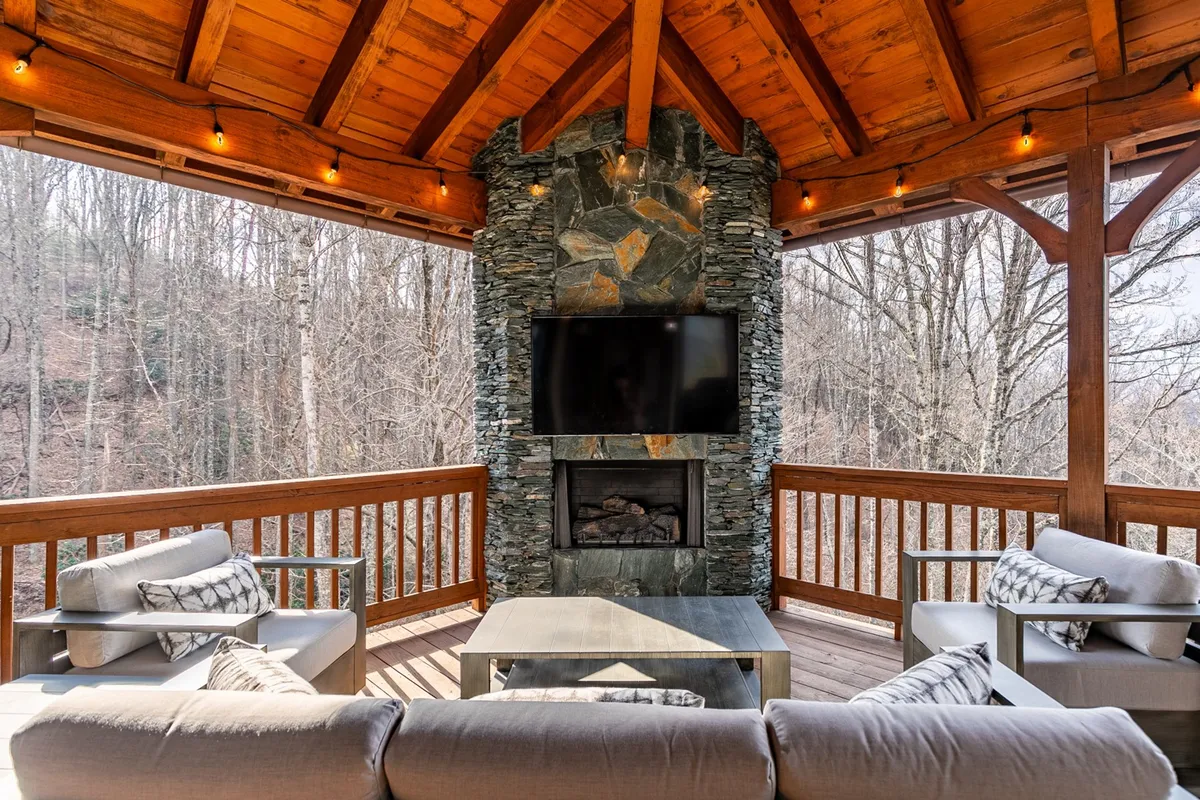 A welcoming covered outdoor deck featuring a prominent stone fireplace, a mounted television, comfortable seating, and exposed wooden beams. The space is arranged for relaxing, socializing, and enjoying the scenic surroundings, with a cozy, warm ambiance ideal for fireside chats or watching sunsets.