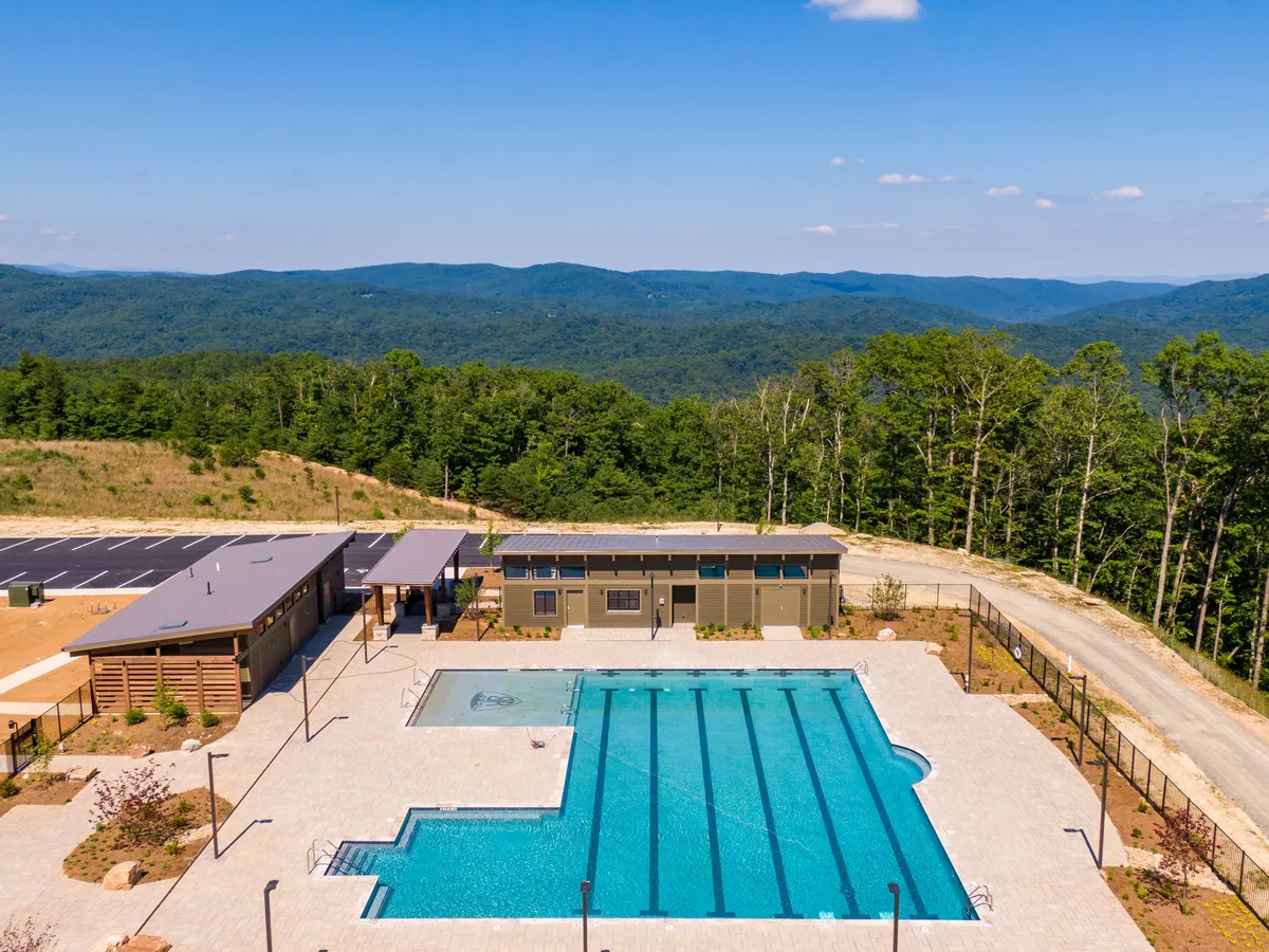 A communal swimming pool set against the backdrop of lush greenery and a community building, illustrating the amenities available within the gated mountain community. The environment looks peaceful and exclusive, with ample space for relaxation and community gatherings.