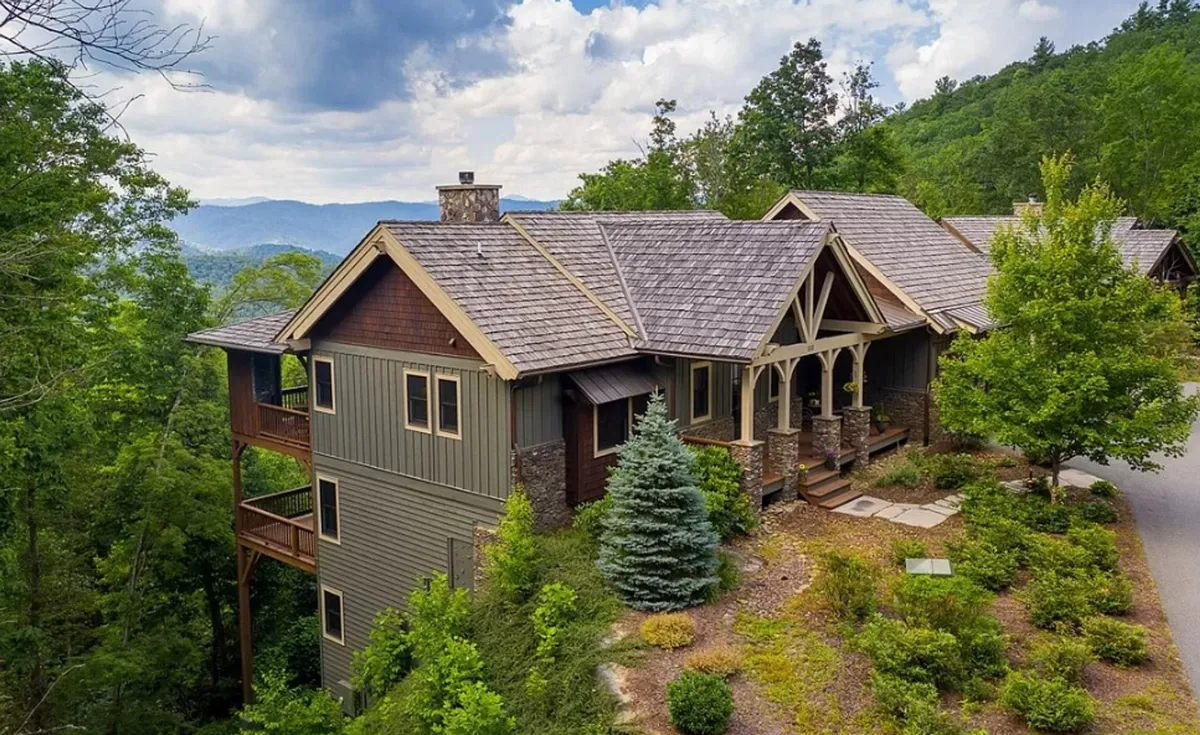 A scenic exterior shot showcasing a rustic porch with sturdy wooden beams, a stone pathway leading up, lush greenery surrounding the space, and a distant view of the mountains. The porch suggests a welcoming place for shared meals or relaxing outdoors while taking in the mountain landscape.